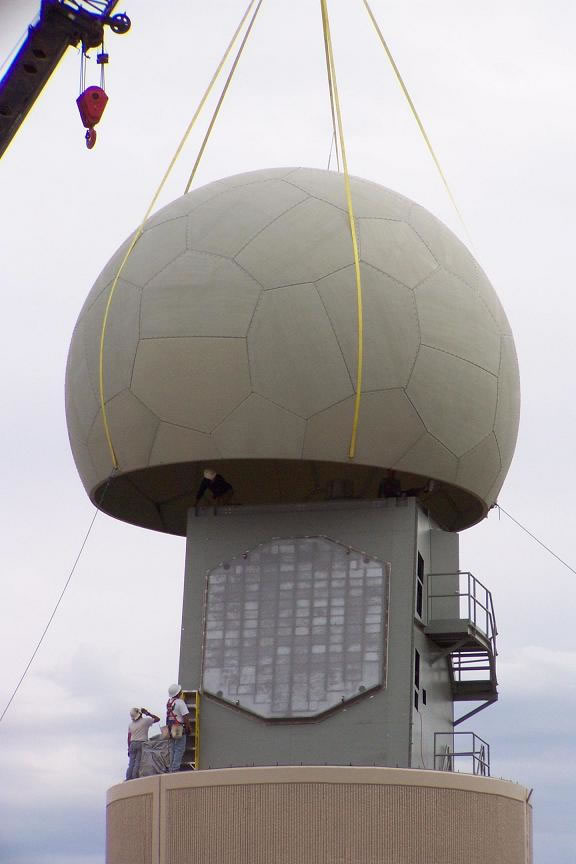 A radar dome for the phased array radar is being lowered by a crane over a flat panel radar anchored to a pedastal.