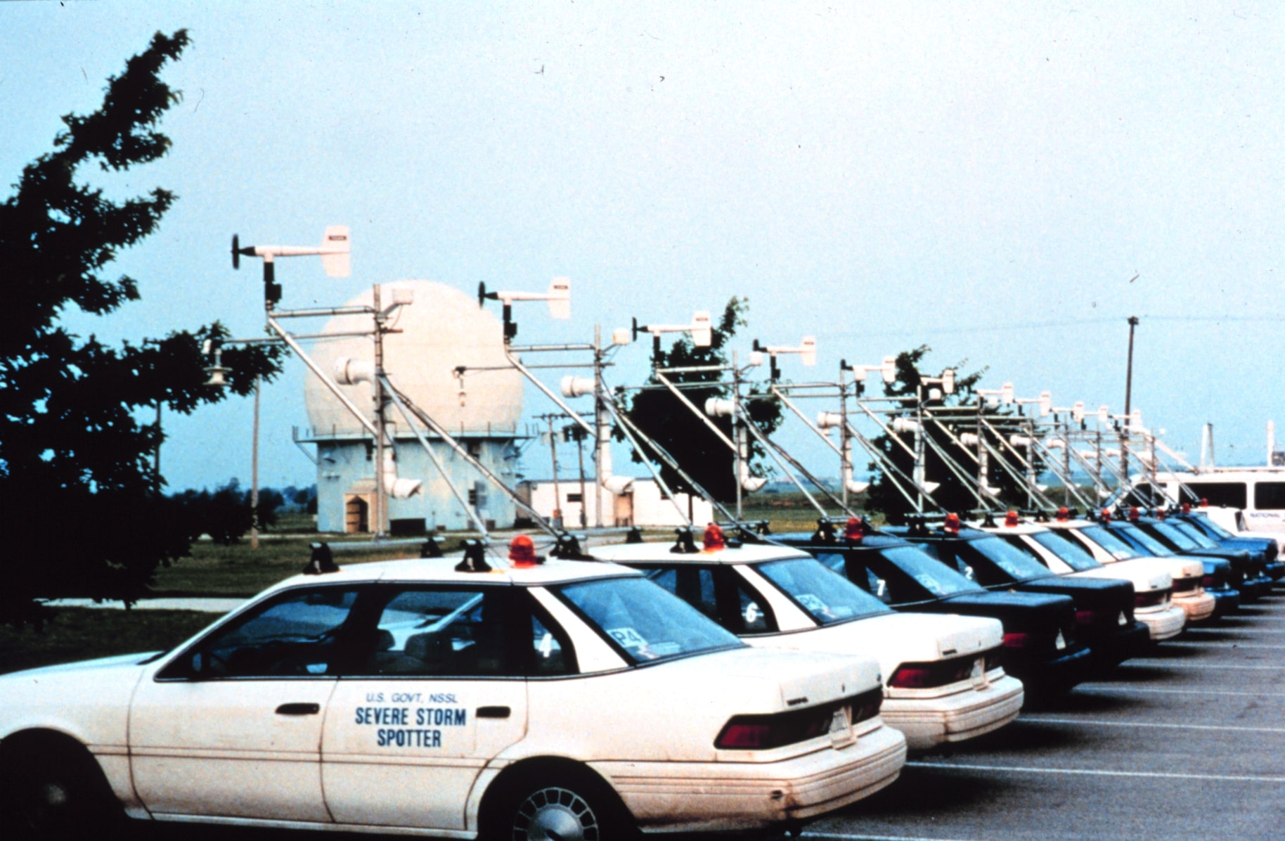 A photo of the original fleet of Mobile Mesonets preparing for the Verification of the Origins of Rotation in Tornadoes EXperiment (VORTEX). Several cars of the same make and model are fixed with weather equipment on top of them and parked in a row, ready for use in the experiment.